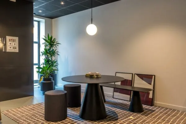 Modern ingerichte zithoek met ronde zwarte tafel, bijpassende krukken, hanglamp en groene planten op een geruite vloer.